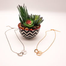 Load image into Gallery viewer, Gold tone and silver tone necklaces with open heart charms intertwined with open pet paw charms.  Displayed on white background with succulent plant as accent. 
