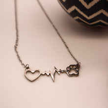 Load image into Gallery viewer, Silver necklace with open heart, heartbeat rhythm, and open pet paw.  Closeup on white background with partial image of pot in background. 
