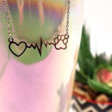 Load image into Gallery viewer, Silver necklace with open heart, heartbeat rhythm, and open pet paw.  Displayed hanging on side of irridescent cup with succulent in background. 
