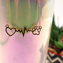 Load image into Gallery viewer, Gold tone necklace with open heart, heartbeat rhythm, and open pet paw. Displayed hanging on side of irridescent cup with succulent in background.
