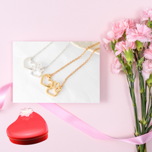 Load image into Gallery viewer, Gold tone and silver tone necklaces with open heart charms intertwined with open pet paw charms.  Displayed on white with pink background, pink flowers and red heart candy tin.  
