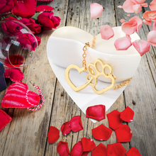 Load image into Gallery viewer, Gold tone necklace with open heart charms intertwined with open pet paw charms.  Displayed hanging over edge of white bowl on background with wood, roses, and flower petals. 
