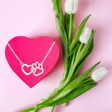 Load image into Gallery viewer, Silver tone necklace with open heart charms intertwined with open pet paw charms.  Displayed laying on pink heart candy box with tulips beside it. 
